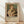 May include: Framed exhibition poster featuring Renoir's 1875 painting of a blonde woman in white dress holding a tabby cat, displayed against white wall on light wooden floor. Poster has cream background with brown text reading "PIERRE AUGUSTE exhibition" and "Woman with a Cat". Natural window light creates soft shadows across the scene.