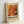 May include: Framed exhibition poster leaning against white wall. Features abstract paint pour artwork in warm orange, red, yellow and black swirls with white splashes. Text reads "POLLOCK" and "EMBRACE THE CHAOTIC REALM OF CREATIVITY" with exhibition dates. Light wood frame on cream background with vintage-style typography.