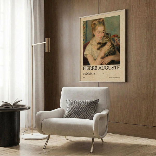 May include: Framed vintage-style art poster featuring Renoir's painting of a blonde woman holding a striped cat, displayed on wood-paneled wall above modern cream armchair with geometric pillow. Room has brass floor lamp, dark round side table with open book, and natural lighting from window with curtains.