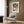 May include: Paul Klee abstract geometric artwork poster in light wood frame, displayed on warm wood panel wall above cream upholstered armchair with patterned pillow. Colorful mosaic-style composition features overlapping squares and rectangles in blues, reds, greens, yellows, and earth tones. Modern reading nook setting with brass floor lamp, dark round side table with open book, and natural lighting from window with sheer curtains.