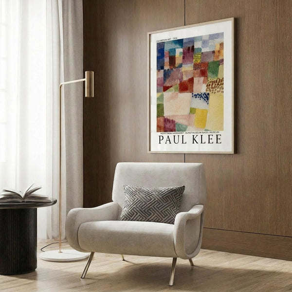 May include: Paul Klee abstract geometric artwork poster in light wood frame, displayed on warm wood panel wall above cream upholstered armchair with patterned pillow. Colorful mosaic-style composition features overlapping squares and rectangles in blues, reds, greens, yellows, and earth tones. Modern reading nook setting with brass floor lamp, dark round side table with open book, and natural lighting from window with sheer curtains.