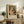May include: Framed art print displaying a woman in white clothing holding a striped tabby cat, with exhibition-style text below reading "PIERRE AUGUSTE exhibition" and "Woman with a Cat 1875". The beige-framed poster leans against a white wall on a rustic wooden console table alongside a fern plant, ceramic vase, stacked books, and table lamp. A beige upholstered chair with brown throw pillow sits nearby in this neutral-toned living space.