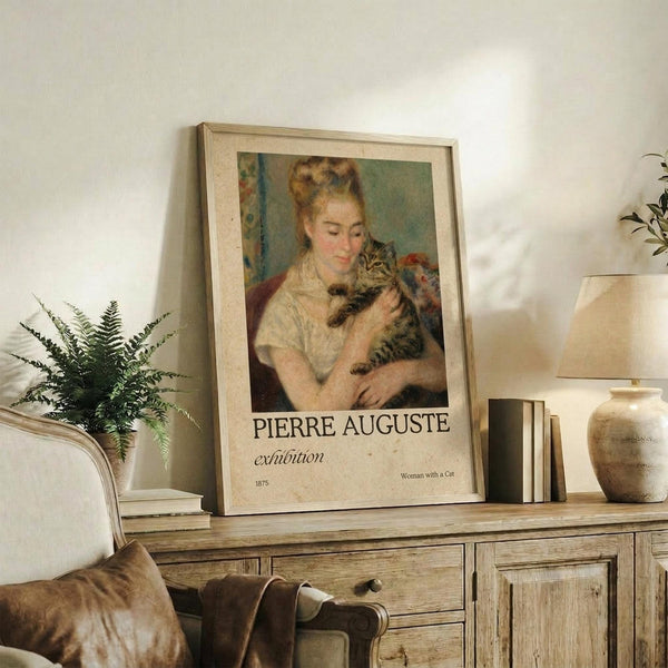 May include: Framed art print displaying a woman in white clothing holding a striped tabby cat, with exhibition-style text below reading "PIERRE AUGUSTE exhibition" and "Woman with a Cat 1875". The beige-framed poster leans against a white wall on a rustic wooden console table alongside a fern plant, ceramic vase, stacked books, and table lamp. A beige upholstered chair with brown throw pillow sits nearby in this neutral-toned living space.