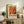 May include: A framed abstract art poster with swirling orange, red, and yellow paint patterns displayed on a rustic wooden sideboard. The artwork features fluid, marbled textures in warm autumn tones with black accents. The poster is propped against a white wall alongside a small fern plant, stacked books, and ceramic pottery, creating a cozy living room vignette with earth-toned furniture and soft natural lighting.