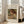 May include: Framed art print of Pierre Auguste Renoir painting showing young child with reddish-brown hair reading a book, rendered in soft impressionist brushstrokes with warm earth tones. White matted frame leans against beige armchair in bright living room with large windows and natural sunlight streaming across hardwood floors.