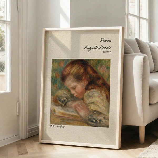 May include: Framed art print of Pierre Auguste Renoir painting showing young child with reddish-brown hair reading a book, rendered in soft impressionist brushstrokes with warm earth tones. White matted frame leans against beige armchair in bright living room with large windows and natural sunlight streaming across hardwood floors.