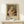 May include: Framed art print displaying a soft impressionist painting of a young woman with upswept auburn hair holding a striped tabby cat. The artwork shows warm peachy and green tones in a classical style. Text below reads "PIERRE AUGUSTE exhibition" and "1875 Woman with a Cat." Natural wood frame sits on light wooden surface against cream wall, with small white wrapped gift nearby.