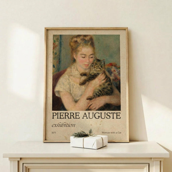 May include: Framed art print displaying a soft impressionist painting of a young woman with upswept auburn hair holding a striped tabby cat. The artwork shows warm peachy and green tones in a classical style. Text below reads "PIERRE AUGUSTE exhibition" and "1875 Woman with a Cat." Natural wood frame sits on light wooden surface against cream wall, with small white wrapped gift nearby.