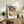 May include: Framed Paul Klee exhibition poster with colorful abstract geometric squares in blues, reds, greens, and yellows, displayed on rustic wooden sideboard alongside potted fern, ceramic vase, and stacked books in neutral-toned living room with beige upholstered chair.