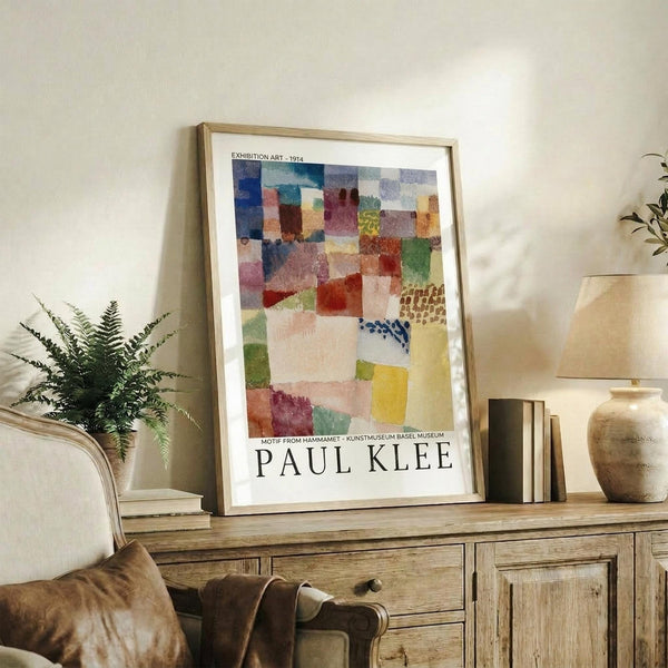 May include: Framed Paul Klee exhibition poster with colorful abstract geometric squares in blues, reds, greens, and yellows, displayed on rustic wooden sideboard alongside potted fern, ceramic vase, and stacked books in neutral-toned living room with beige upholstered chair.
