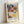 May include: A framed reproduction poster featuring Paul Klee's abstract watercolor artwork from 1914. The composition shows overlapping geometric shapes in soft blues, greens, yellows, pinks, and earth tones with translucent washes of color. Text at top reads "Exhibition Art - 1914" and bottom shows "Motif from Hammamet - Kunstmuseum Basel Museum" with the artist's name. The poster is displayed in a light wood frame leaning against a white wall.