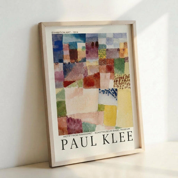 May include: A framed reproduction poster featuring Paul Klee's abstract watercolor artwork from 1914. The composition shows overlapping geometric shapes in soft blues, greens, yellows, pinks, and earth tones with translucent washes of color. Text at top reads "Exhibition Art - 1914" and bottom shows "Motif from Hammamet - Kunstmuseum Basel Museum" with the artist's name. The poster is displayed in a light wood frame leaning against a white wall.