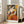 May include: Framed geometric abstract art print featuring a seated woman in cubist style with angular shapes in orange, yellow, red, navy blue, and cream colors. The poster is displayed in a light wood frame leaning against white walls near large windows, positioned next to a beige upholstered sofa in a bright modern living room with hardwood floors.
