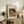 May include: Framed reproduction of Renoir's "Child Reading" showing a young girl with reddish hair reading a book, displayed on rustic wooden sideboard alongside fern plant, ceramic vase, and stacked books in neutral-toned living room setting with cream upholstered chair visible.