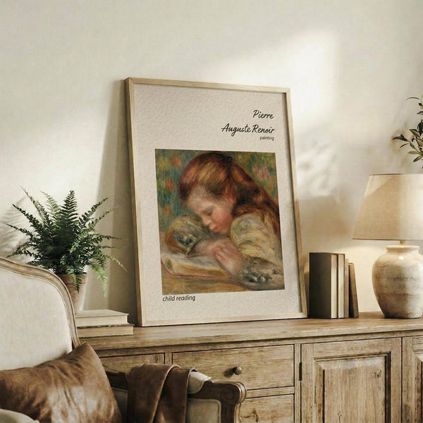 May include: Framed reproduction of Renoir's "Child Reading" showing a young girl with reddish hair reading a book, displayed on rustic wooden sideboard alongside fern plant, ceramic vase, and stacked books in neutral-toned living room setting with cream upholstered chair visible.