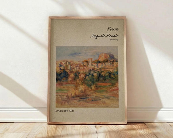 May include: Framed poster print featuring a Renoir landscape painting with warm earth tones showing buildings, trees, and countryside. The artwork displays impressionist brushwork in oranges, greens, and browns. Housed in a light wood frame with beige matting, the print leans against a white wall on light wooden flooring with soft natural lighting creating diagonal shadows.