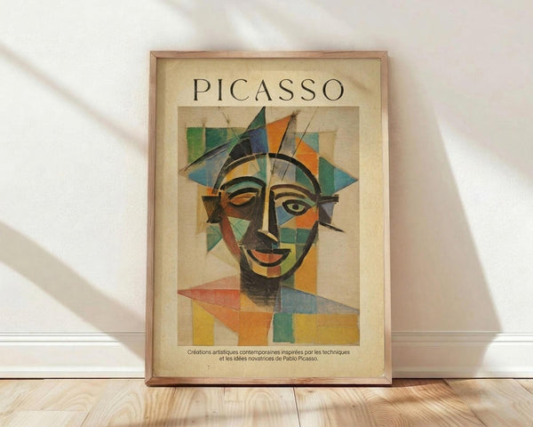 May include: Framed poster print featuring a colorful cubist-style portrait with geometric facial features in blues, oranges, greens, and yellows. The word "PICASSO" appears at the top with French text below. Natural wood frame with cream matting, displayed against white wall on light wooden floor with soft window light shadows.