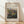 May include: Framed art print of an Impressionist landscape painting showing rolling hills, lush vegetation, and mountains in soft greens, blues, and earth tones. The wooden frame sits against a white wall on light wood flooring with natural sunlight casting diagonal shadows across the scene.
