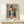 May include: Framed Picasso-style cubist portrait of woman wearing geometric hat in orange, black, and white tones, displayed on light wood surface with small wrapped gift, neutral wall background, natural lighting creating soft shadows