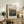 May include: Framed Renoir landscape print displayed on rustic wooden sideboard next to fern plant, books, and ceramic vase. The artwork shows rolling green hills and countryside in impressionist style with soft brushstrokes. Natural lighting creates warm ambiance in cream-walled room with beige upholstered chair visible in corner.