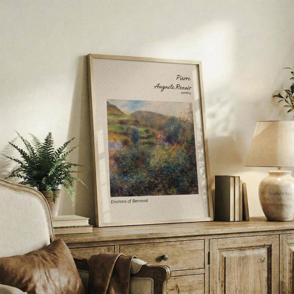 May include: Framed Renoir landscape print displayed on rustic wooden sideboard next to fern plant, books, and ceramic vase. The artwork shows rolling green hills and countryside in impressionist style with soft brushstrokes. Natural lighting creates warm ambiance in cream-walled room with beige upholstered chair visible in corner.