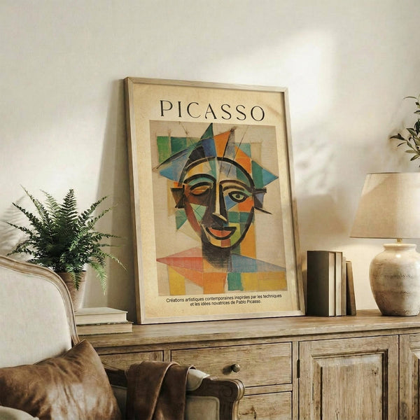May include: Framed Picasso-style cubist portrait poster with geometric colorful face design displayed on rustic wooden sideboard alongside fern plant, books, ceramic vase, and beige armchair in modern living room setting with neutral walls
