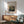 May include: Framed art print featuring Pierre Auguste Renoir's impressionist landscape painting of rolling green hills and countryside around Berneval. The poster has a beige mat with brown wooden frame, displayed above a mid-century modern wood credenza with white ceramic lamp. A mint green upholstered armchair with wooden arms sits nearby on hardwood floors. Soft natural sunlight streams across the white wall, creating geometric shadows in this minimalist living room setting.