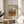May include: Framed art print displaying an impressionist landscape painting with warm earth tones showing Mediterranean-style buildings and trees. The cream-colored poster features black text identifying the artist and date. Displayed on a light wooden round side table next to a beige upholstered chair, with a plant's leaves visible in soft natural lighting creating shadows on white walls.