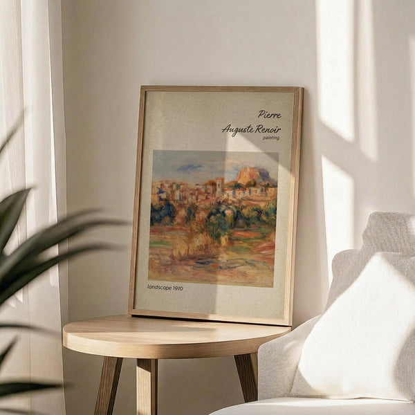 May include: Framed art print displaying an impressionist landscape painting with warm earth tones showing Mediterranean-style buildings and trees. The cream-colored poster features black text identifying the artist and date. Displayed on a light wooden round side table next to a beige upholstered chair, with a plant's leaves visible in soft natural lighting creating shadows on white walls.