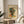 May include: Framed Picasso-style cubist portrait poster with geometric colorful face design in yellows, blues, greens and oranges, displayed on light wood round side table next to beige upholstered chair with knitted throw pillow. Natural sunlight streams across white walls creating soft shadows, with green plant leaves visible in corner of modern minimalist living room setting.