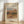 May include: A framed reproduction print of a Pierre Auguste Renoir landscape painting from 1910, displayed in a light wooden frame against a white wall. The impressionist artwork shows a Mediterranean hillside village with warm earth tones, buildings nestled among trees, and soft brushstrokes typical of Renoir's style. The print features cream matting with elegant typography identifying the artist and date.