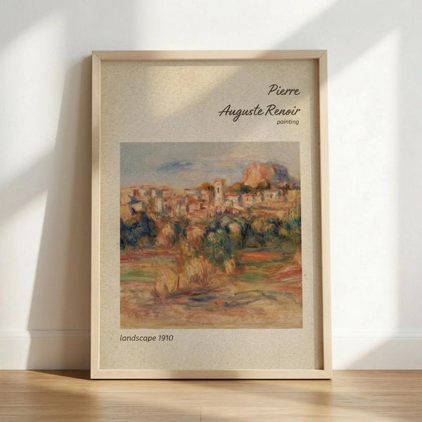 May include: A framed reproduction print of a Pierre Auguste Renoir landscape painting from 1910, displayed in a light wooden frame against a white wall. The impressionist artwork shows a Mediterranean hillside village with warm earth tones, buildings nestled among trees, and soft brushstrokes typical of Renoir's style. The print features cream matting with elegant typography identifying the artist and date.