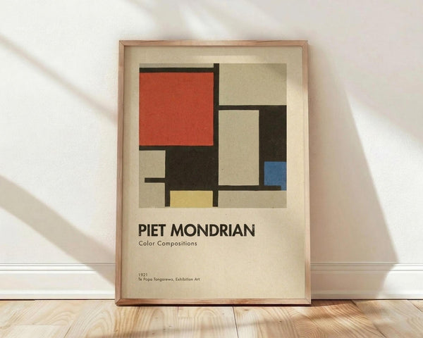 May include: Framed exhibition poster featuring a geometric abstract composition with red, blue, yellow, black and beige rectangular shapes in Mondrian's distinctive style. The cream-colored poster displays black text below the artwork. Shown in a light wooden frame leaning against a white wall on wooden flooring with soft natural lighting creating diagonal shadows.