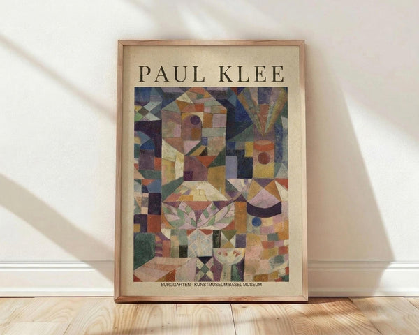 May include: Framed Paul Klee abstract art print leaning against white wall on light wooden floor. The colorful geometric artwork features mosaic-like squares and shapes in blues, greens, oranges, purples and earth tones, creating fragmented architectural forms. Natural wood frame with cream matting. Soft window light creates diagonal shadows across the white textured wall behind the medium-sized poster.