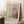 May include: Framed Paul Klee exhibition poster leaning against white wall in cozy room setting. The artwork features abstract floral design with dotted background in soft pastels - pink circles, green stem, and colorful speckled pattern. Light wood frame complements the delicate colors. White coffee cup sits on wooden block nearby, with cream knit blanket draped beside the poster on white fluffy rug.