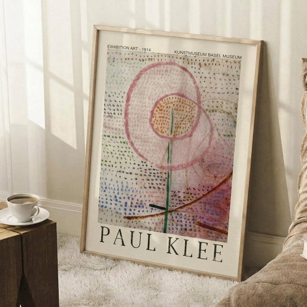May include: Framed Paul Klee exhibition poster leaning against white wall in cozy room setting. The artwork features abstract floral design with dotted background in soft pastels - pink circles, green stem, and colorful speckled pattern. Light wood frame complements the delicate colors. White coffee cup sits on wooden block nearby, with cream knit blanket draped beside the poster on white fluffy rug.