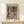 May include: Framed poster print of abstract geometric artwork featuring colorful overlapping shapes, cubes, and angular forms in muted tones of purple, green, yellow, orange and pink. The composition suggests fragmented architectural or garden elements rendered in a cubist style. Wood frame displays on light wooden surface alongside small wrapped gift box with twine and greenery accent against cream colored wall.