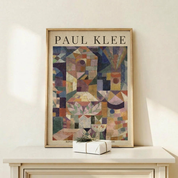 May include: Framed poster print of abstract geometric artwork featuring colorful overlapping shapes, cubes, and angular forms in muted tones of purple, green, yellow, orange and pink. The composition suggests fragmented architectural or garden elements rendered in a cubist style. Wood frame displays on light wooden surface alongside small wrapped gift box with twine and greenery accent against cream colored wall.