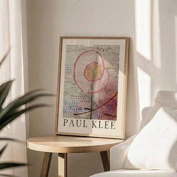 May include: Framed Paul Klee exhibition poster featuring abstract floral artwork with pink swirled petals, dotted background in blues and earth tones, and green stem. Light wood frame sits on round wooden side table next to beige upholstered chair. Soft natural lighting creates shadows on white wall. Plant leaves visible in corner of bright, minimalist room setting.