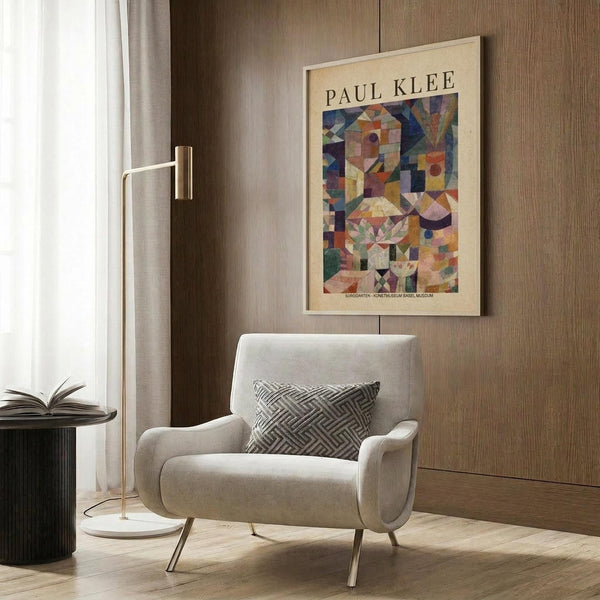 May include: Paul Klee abstract geometric art print in light wooden frame hanging on warm wood paneled wall. Colorful cubist composition with overlapping shapes in blues, oranges, purples, and earth tones. Styled in modern reading nook with cream upholstered armchair, geometric patterned pillow, brass floor lamp, and dark round side table with open book.