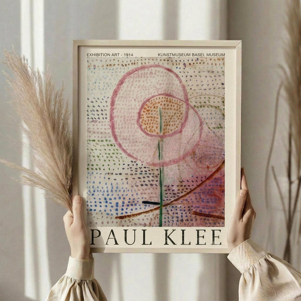 May include: Person holding framed art poster featuring abstract botanical design with pink circular forms, green stem, and colorful dotted pattern background. Light wood frame against white curtained window, dried wheat stems visible on left side.