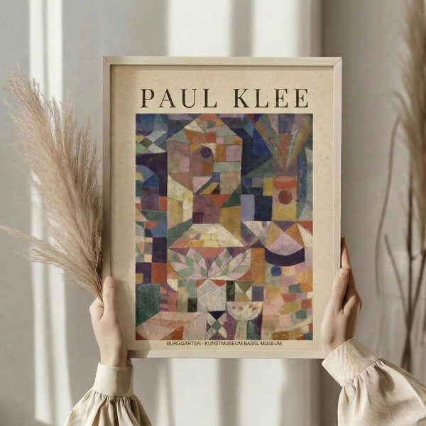 May include: Person in cream ruffled sleeves holding framed Paul Klee art print featuring abstract geometric composition in colorful squares, triangles and botanical motifs. Light wood frame against soft neutral background with dried pampas grass stems visible on left side.