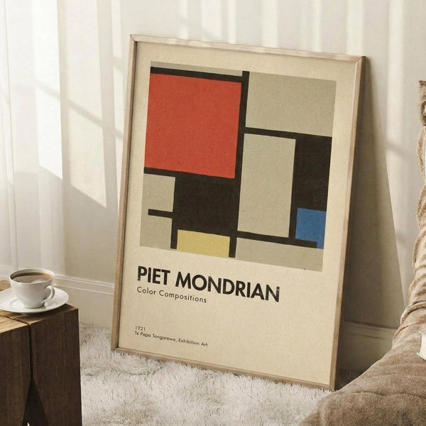 May include: Framed geometric art print leaning against white wall corner on textured cream rug. Abstract composition features red, blue, yellow, black, and beige rectangular blocks separated by thick black lines. Light wood frame, exhibition-style text below artwork. Coffee cup on wooden block nearby, soft natural lighting creates window shadows on wall.