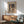 May include: Framed Paul Klee art print hanging on white wall above wooden sideboard with white ceramic lamp. Mid-century modern sage green upholstered armchair with wooden frame sits nearby. The colorful abstract artwork features geometric shapes in earth tones - browns, greens, oranges, and blues arranged in cubist style. Natural sunlight creates shadows across the minimalist room setting with light wood furniture and neutral decor.