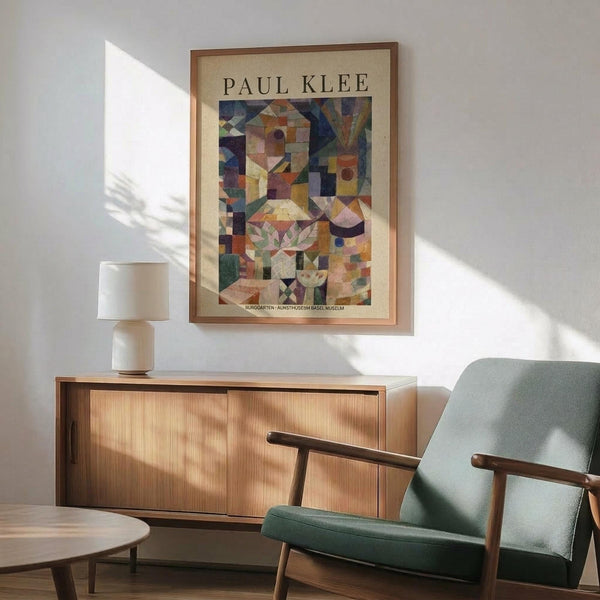 May include: Framed Paul Klee art print hanging on white wall above wooden sideboard with white ceramic lamp. Mid-century modern sage green upholstered armchair with wooden frame sits nearby. The colorful abstract artwork features geometric shapes in earth tones - browns, greens, oranges, and blues arranged in cubist style. Natural sunlight creates shadows across the minimalist room setting with light wood furniture and neutral decor.