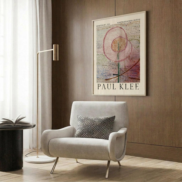May include: Paul Klee exhibition poster displayed in modern reading nook with beige upholstered armchair, geometric patterned pillow, brass floor lamp, and dark round side table with open book. The framed artwork features abstract circular forms in pink and orange tones with dotted patterns against wood-paneled wall. Soft natural lighting from nearby window illuminates the cozy corner setup on hardwood flooring.
