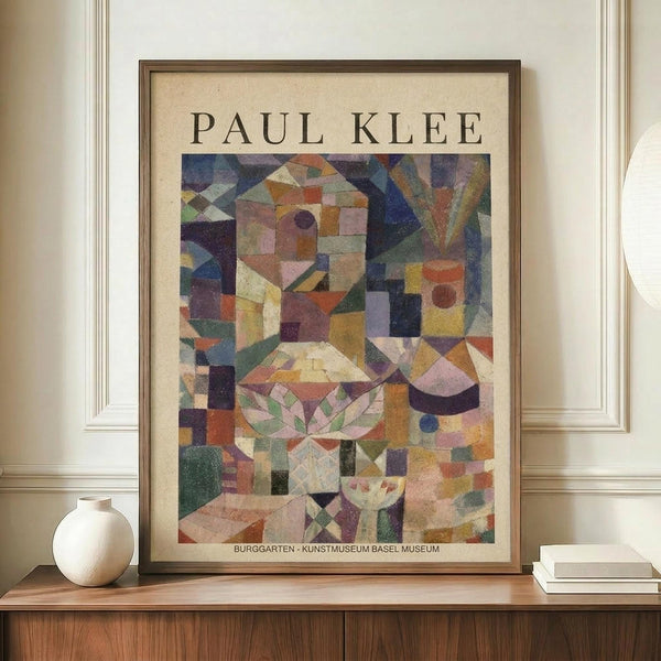 May include: Framed Paul Klee abstract art print displayed on wooden sideboard against cream wall with white ceramic vase and stacked books. The colorful geometric artwork features fragmented architectural forms in muted blues, oranges, purples and greens with botanical motifs. Museum exhibition poster in natural wood frame on elegant furniture piece.