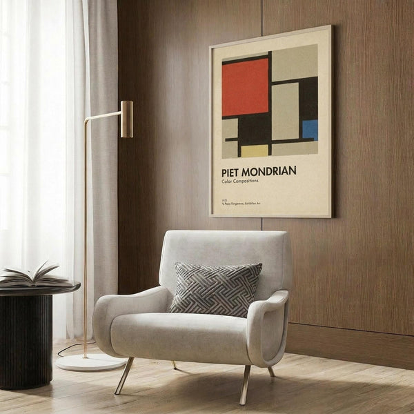 May include: Framed Piet Mondrian-style geometric art print with red, blue, yellow and black rectangular shapes on cream background, displayed above a beige upholstered armchair with patterned pillow. Modern living room setting features brass floor lamp, dark wood paneled wall, black side table with open book, and neutral curtains.