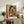 May include: Framed cubist-style artwork showing two embracing women with geometric faces in warm oranges, blues, and reds, displayed on a rustic wood sideboard alongside fern plant, books, ceramic vase, and cream upholstered chair in neutral-toned living room setting.
