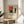 May include: A framed poster featuring Piet Mondrian's geometric abstract art with colorful rectangles in red, blue, yellow, and beige separated by bold black lines. The print sits on a light wood round side table next to a beige upholstered chair with plant leaves visible in soft natural lighting streaming through white curtains.